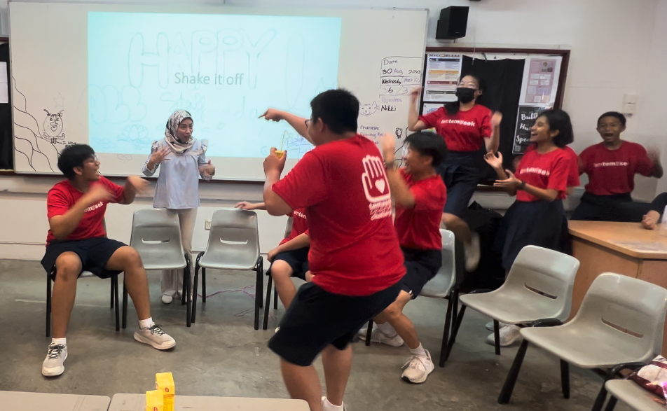Students in red shirts dance energetically in a classroom with a projector showing "Shake it off."